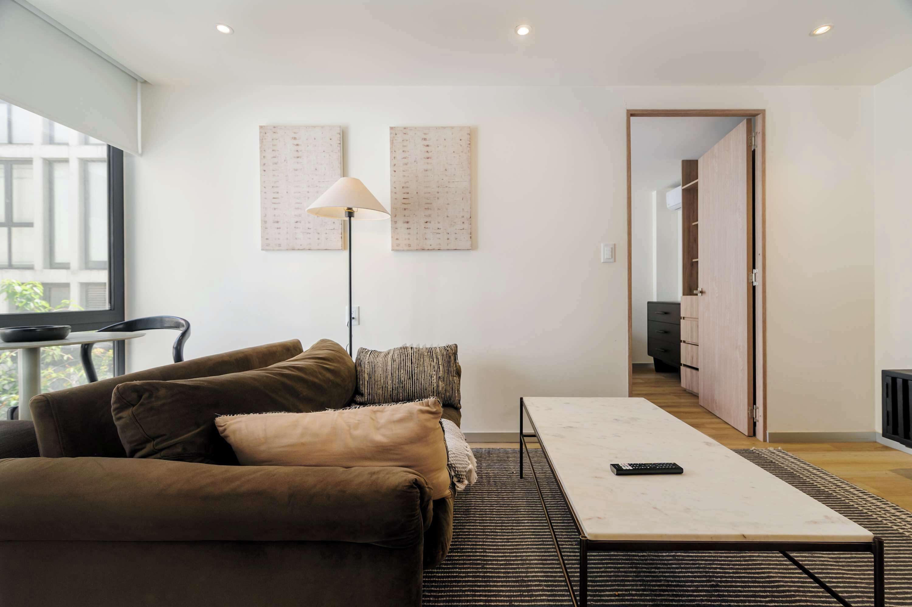 Een moderne woonkamer met een bank, een salontafel en een vloerlamp. Er is een deur die leidt naar een andere kamer en een raam zorgt voor natuurlijk licht.