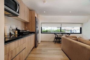 A modern kitchen with wooden cabinets, a black countertop, and a built-in microwave. There is a window with a view of buildings and greenery. - w* | Luxury 1BR in Cuauhtemoc w/ AC (Ciudad de México)