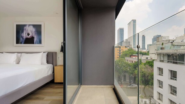 Une chambre moderne avec un lit, une œuvre d’art encadrée et une vue sur la ville depuis un balcon.