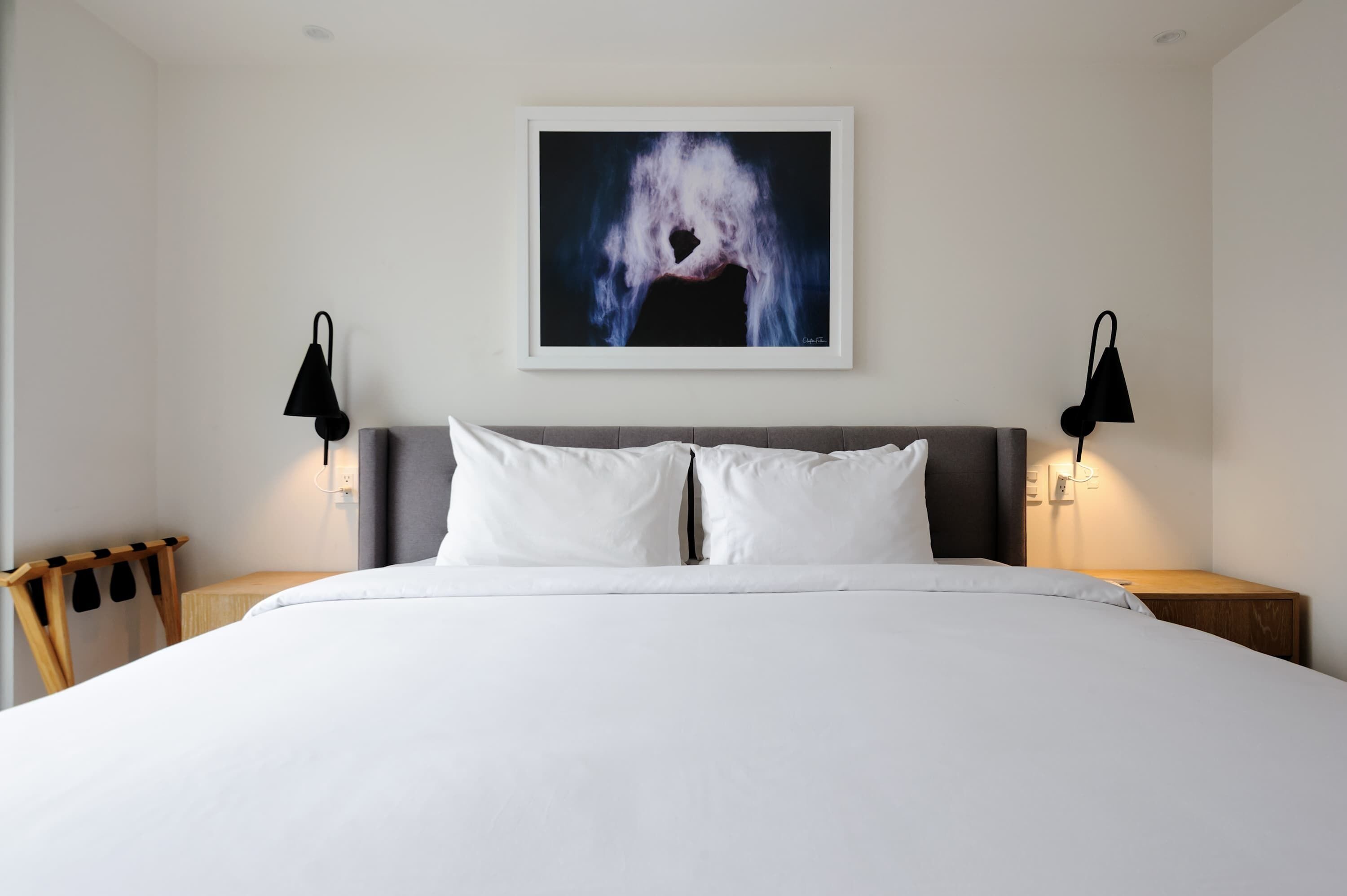 A hotel room with a large bed, two bedside lamps, a framed artwork above the bed, and a wooden nightstand with a lamp.