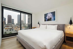 Ein modernes Hotelzimmer mit einem großen Bett, Stadtblick durch raumhohe Fenster, einem gerahmten Kunstwerk an der Wand und einem Nachttisch mit Lampe.