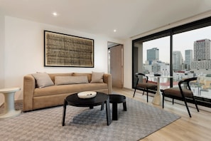 Ein modernes Wohnzimmer mit einer Couch, zwei Tischen, Stühlen, einem großen Fenster mit Blick auf die Stadt und einem gerahmten Kunstwerk an der Wand.
