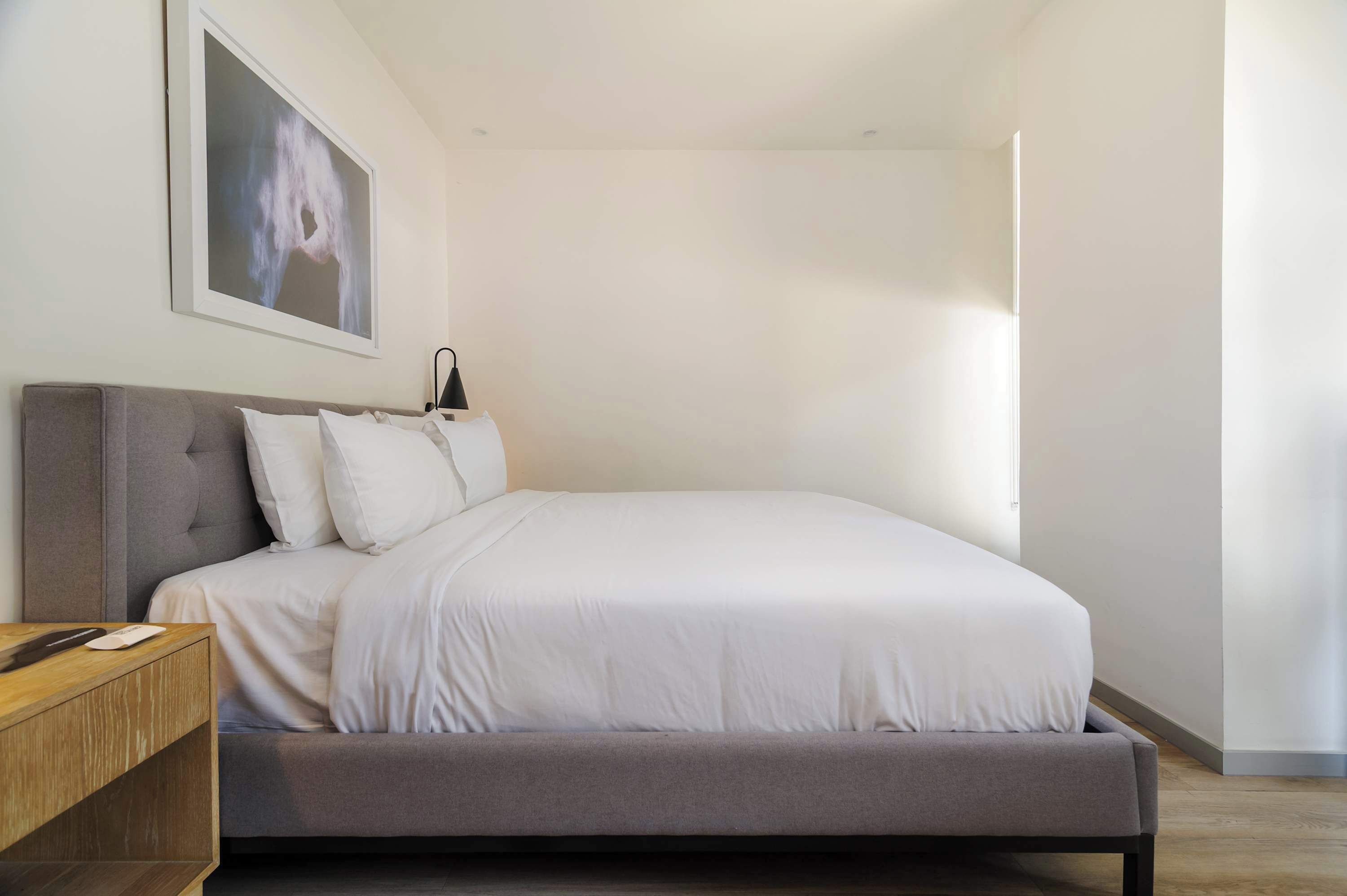 A modern bedroom with a grey upholstered bed, white bedding, a wooden nightstand, and a black desk lamp.