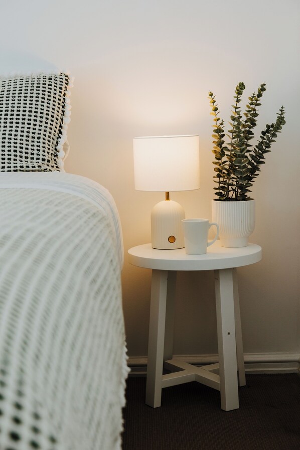 A bed with a white textured blanket, a bedside table with a white lamp, a white mug, and a potted plant. - Bluff Point Coastal Townhouse - 3 Bedrooms - Sleeps 6 - Double Garage (Bluff Point)