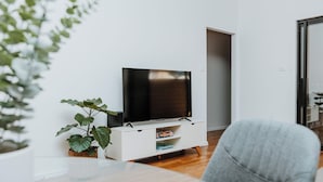 Ein modernes Wohnzimmer mit einem Flachbildfernseher auf einem weißen Schrank, einem grauen Sofa und einer Topfpflanze.