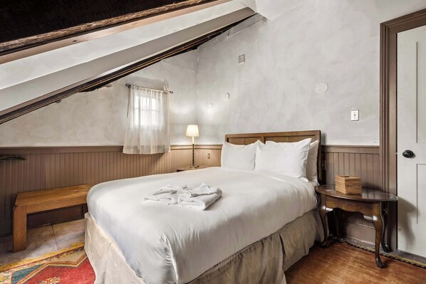 Quarto com cama grande, mesa de cabeceira, um banco e janela com cortina.
