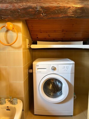 Een compacte wasmachine in een kleine bijkeuken met betegelde wanden, een houten plafond en een wasbak met kranen.