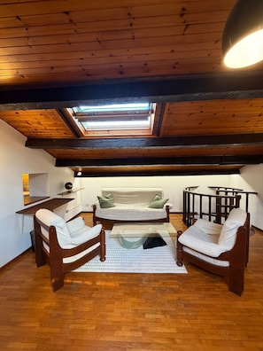 Een slaapkamer in loftstijl met dakraam, houten balken, een bank, twee fauteuils, een glazen salontafel en een klein bureau.