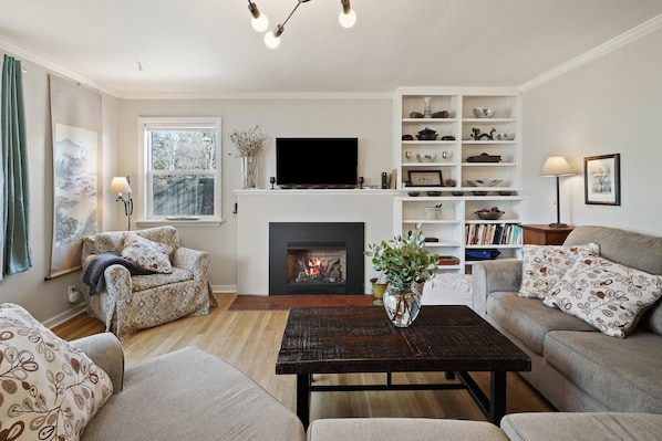 A living room with a fireplace, sofa, armchair, coffee table, and a TV. - NW Nest 30 Day Minimum (Bend)