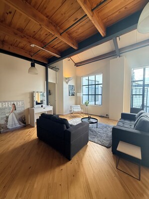 A spacious living room with wooden floors, a black sofa, a white chair, a small round table, and a large window. - Stylish New York-Style Loft in Parnell  Spacious Auckland Escape with parking. (parnell)