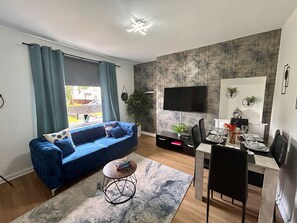 Un soggiorno moderno con un divano blu, un tavolo da pranzo con sedie, una TV a parete e un'ampia finestra con tende.