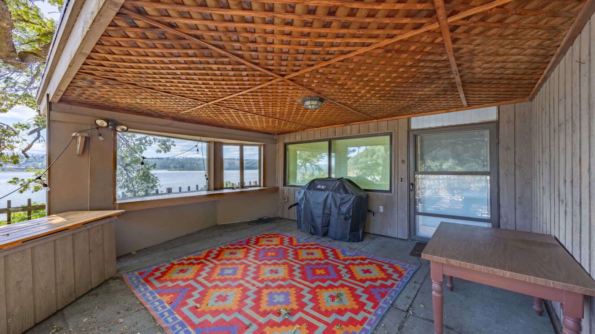 Un espacio al aire libre techado, con techo de madera, alfombra colorida, una parrilla y una mesa.