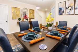Comedor con una mesa larga dispuesta para seis personas, con platos azules, cubiertos dorados y servilletas naranjas.