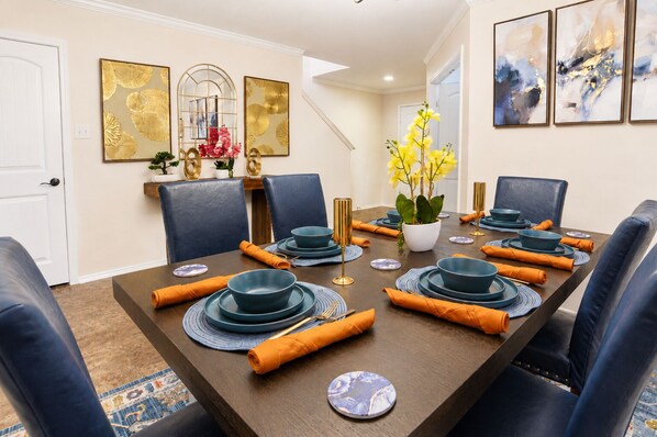 A dining room with a long table set for six, featuring blue plates, gold cutlery, and orange napkins. - Spacious North Austin–Pflugerville Retreat - 6 King Beds - Modern Comfort (Pflugerville)