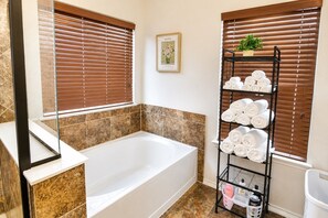 A bathroom with a bathtub, brown tiled walls, a towel rack with rolled towels, and a framed picture on the wall. - Spacious North Austin–Pflugerville Retreat - 6 King Beds - Modern Comfort (Pflugerville)