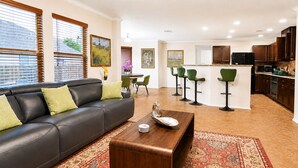 Een woonkamer met een bank, salontafel en groene stoelen. Er is een keuken met kasten en een koelkast.
