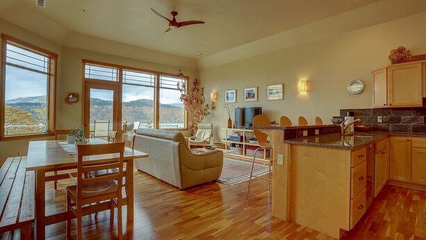 A spacious living area with a wooden dining table, a sofa, and a kitchen with wooden cabinets and a view of the outdoors. - Three Rivers 30 day minimum (Hood River)
