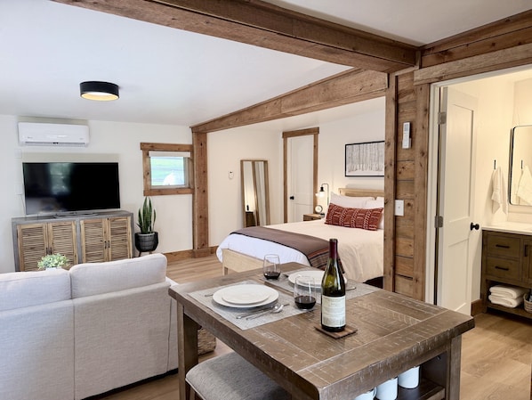 A room with a bed, a TV, a dining table with wine, and a wooden ceiling.