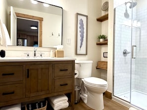 A bathroom with a wooden vanity, a toilet, a shower, and a framed artwork on the wall.