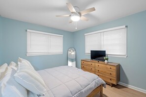 Ein Schlafzimmer mit einem Bett, einem Schminktisch, einem Deckenventilator und zwei Fenstern mit Jalousien.