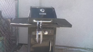 Een zwarte buitenbarbecue met een deksel, handgreep en bedieningsknop, voor een betonnen muur met een hek van gaasdraad.