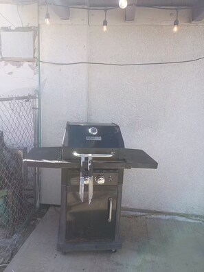 A black outdoor grill with a front lid, a handle, and a control knob, set against a concrete wall with a chain-link fence in the background. - Lovely 1-bedroom house with AC in charming Twentynine Palms (Twentynine Palms)