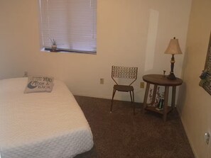 A bedroom with a bed, a chair, a small table with a lamp, a window with blinds, and a wall-mounted shelf. - Lovely 1-bedroom house with AC in charming Twentynine Palms (Twentynine Palms)