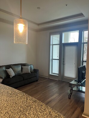 Uma sala de estar com um sofá escuro, uma mesa de centro de vidro, uma luminária pendente e uma janela grande com persianas.