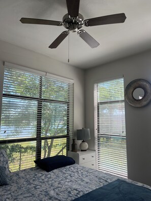Quarto com cama, ventoinha no teto, luminária e janelas amplas com persianas.