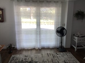 A room with a large window covered by sheer curtains, a standing fan, a patterned rug, and a small table with items on it. - Soothing 1-bedroom cottage with AC in charming Clyde.  Located on quiet 5 acres (Clyde)