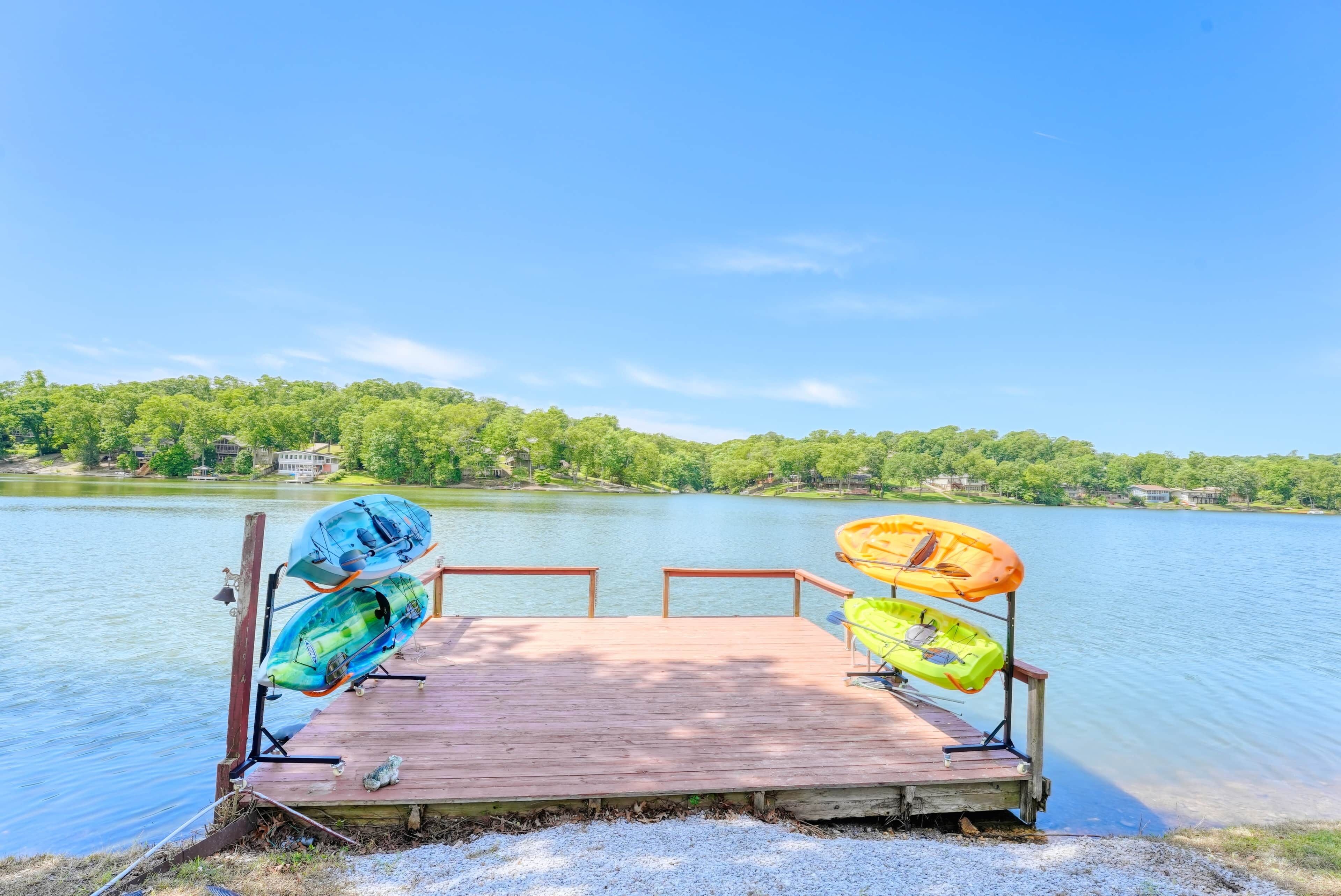 A wooden dock with kayaks and a canoe, overlooking a calm lake with trees and buildings in the background.