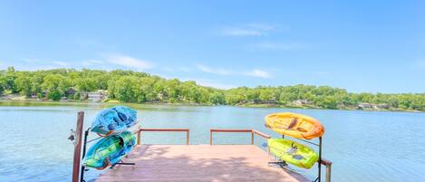 A wooden dock with kayaks and a canoe, overlooking a calm lake with trees and buildings in the background.