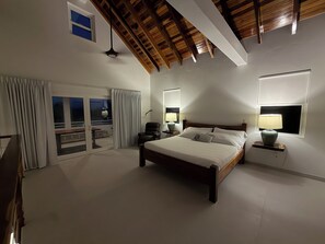 Une chambre dotée d’un grand lit, de deux lampes de chevet, d’un ventilateur de plafond et offrant une vue sur l’extérieur grâce à une porte-fenêtre coulissante qui donne sur un espace extérieur.