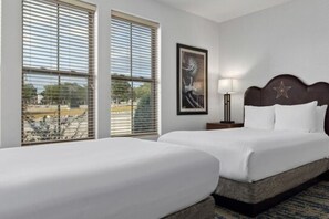 A hotel room with two beds, a framed artwork on the wall, a nightstand with a lamp, and large windows with blinds. - BLOCK WM Stablewood Springs 2br (Ingram)