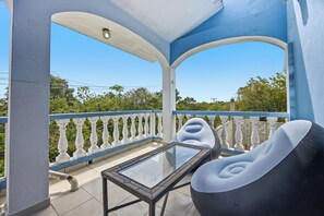 Een balkon met een glazen tafel en twee stoelen, een witte reling, en uitzicht op groen.
