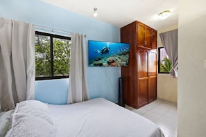 Un dormitorio con cama, televisor en la pared, armario y ventana con cortinas.