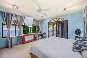 Un dormitorio con cama, escritorio, silla, ventilador de techo y vista a una cascada a través de una ventana.