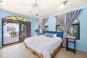 Een slaapkamer met een bed, een plafondventilator, een raam met gordijnen, een nachtkastje en uitzicht op een buitenruimte.