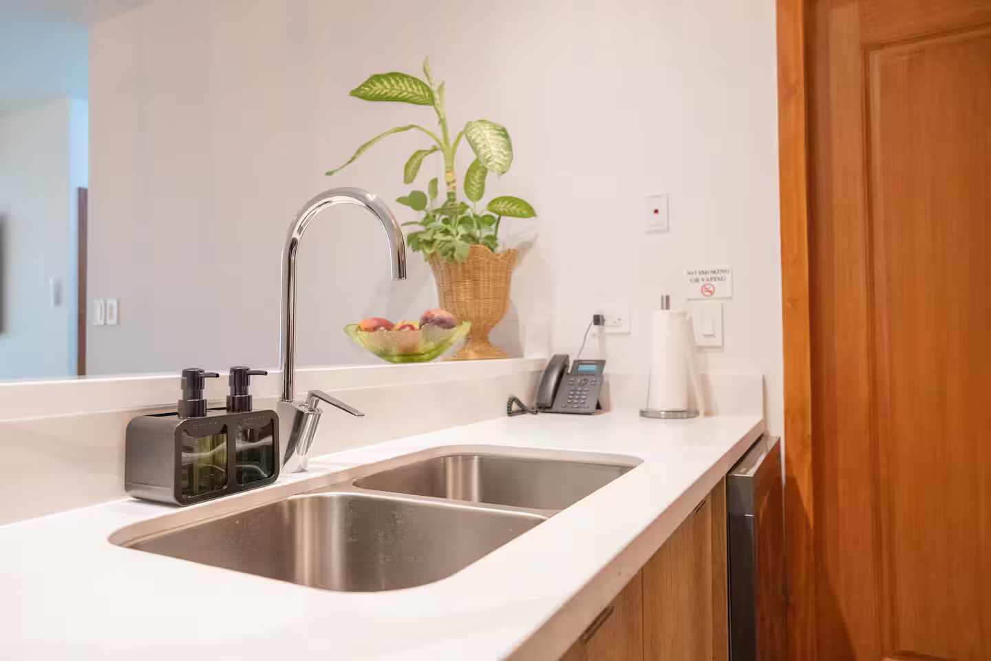 A modern kitchen with a double sink, a plant, a fruit bowl, a phone, and a sign indicating no smoking.