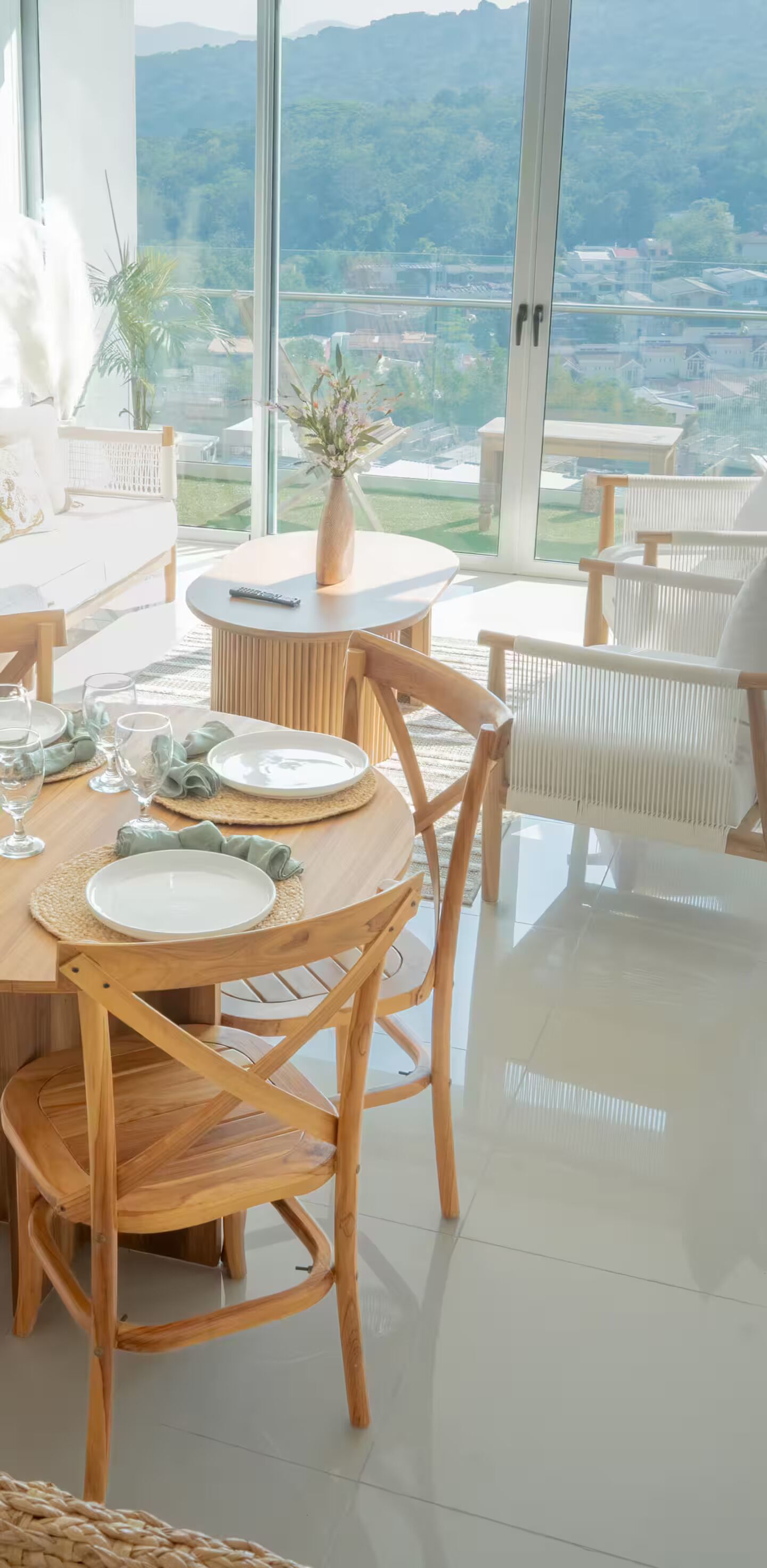 A dining area with a table set for four, surrounded by wicker chairs, with a view of a green landscape through large windows.