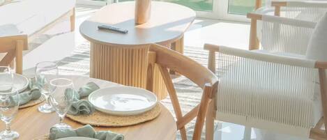 A dining area with a table set for four, surrounded by wicker chairs, with a view of a green landscape through large windows.