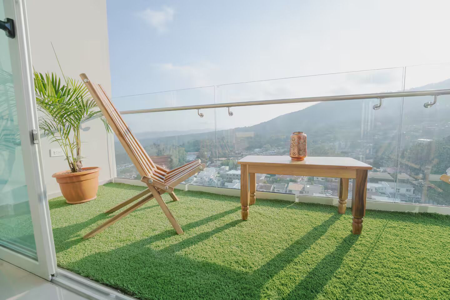 Un balcón con césped artificial, una silla de madera, una mesita y una planta en maceta.