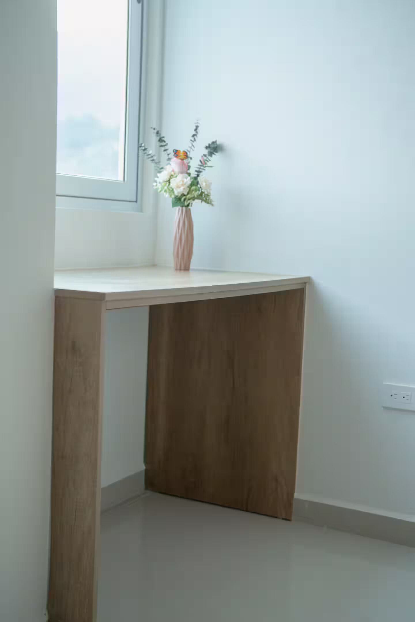 Un bureau d’angle agrémenté d’un vase de fleurs, une fenêtre et un mur blanc.