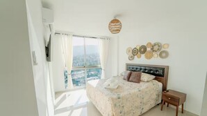 Quarto com cama grande, mesa de cabeceira, TV na parede e vista da cidade através de grandes janelas.
