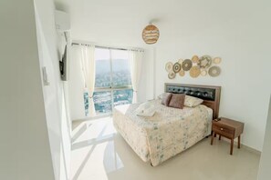 Ein Schlafzimmer mit einem großen Bett, einem Nachttisch, einem an der Wand montierten Fernseher und Blick auf die Stadt durch große Fenster.