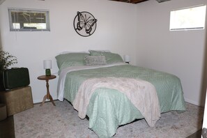 A bedroom with a bed, two bedside lamps, a wall-mounted butterfly decoration, a window with blinds, a small round table with a lamp, and a green suitcase on a bench. - Charming 4 Bedroom Home in Waucoma, Iowa-River front property (Waucoma)