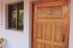 Una puerta de madera con una placa de número, una ventana con cortina y una silla afuera.
