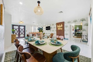 A dining area with a long table set for four, surrounded by chairs, with a television mounted on the wall and a decorative wall panel. - Mins to Beach • Backyard Oasis • Heated Pool & Spa (West Palm Beach)