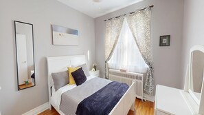 Una camera da letto con un letto, un comodino, uno specchio, un termosifone, una finestra con tende e un quadro appeso al muro.