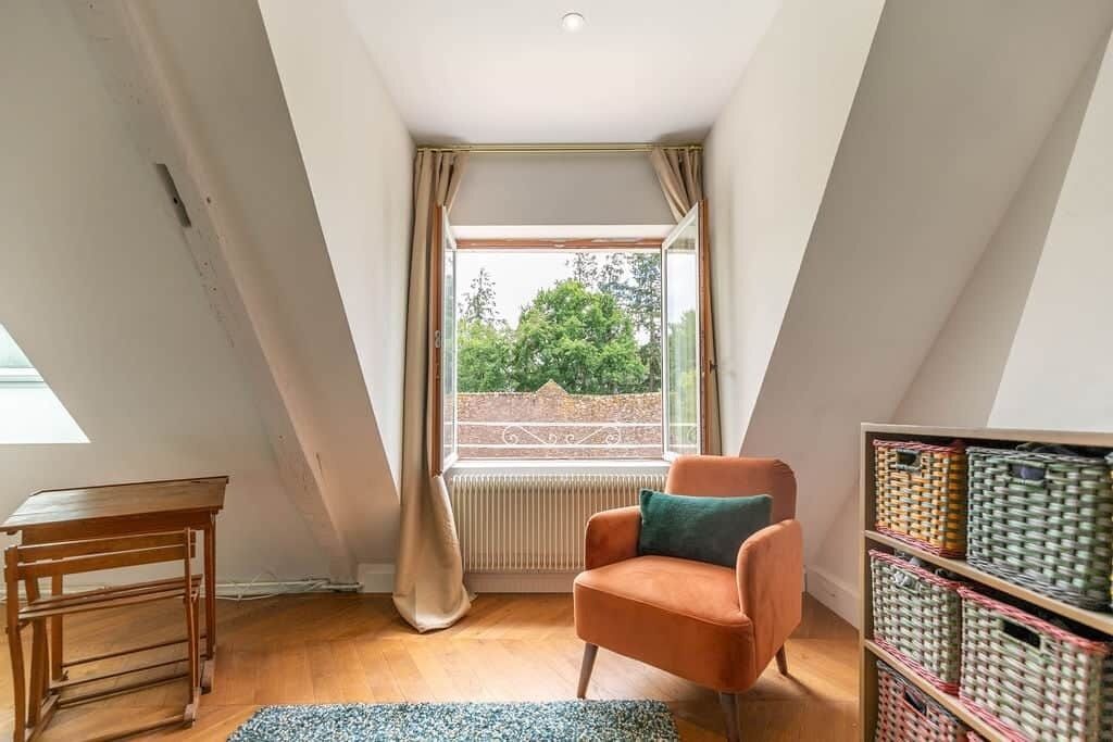 Ein Zimmer mit schrägem Dach, einem mit Vorhängen versehenen Fenster, einem Holztisch, einem orangefarbenen Sessel und Korbwäschekörben.